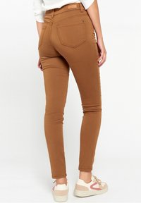 Brown, fitted denim jeans with a smooth texture, five pockets, and subtle stitching details. Paired with light-colored casual shoes.