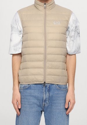 Gilet sans manches matelassé beige avec fermeture éclair devant, porté sur un t-shirt blanc et gris à manches courtes et un jean en denim bleu.
