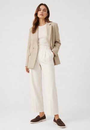 Frau trägt beige Blazer, weißes Oberteil, weiße, hoch taillierte, weit ausgestellte Hose und braune Loafers, steht vor einfarbigem Hintergrund.