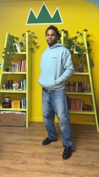 Šviesiai mėlynas džemperis su kapisonu ir juodu logotipu, laisvo kirpimo džinsai ir juodi batai. Lentynos su knygomis ir augalais prie geltonos sienos su žaliais akcentais.