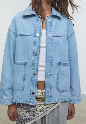 Vrouw draagt een lichtblauwe spijkerjas met metalen knopen over een witte cropped top en een sjaal met bloemenpatroon gestrikt rond een grijze broek.