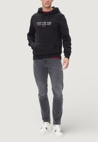 Felpa nera con cappuccio e tasca a marsupio, con logo bianco, abbinata a jeans grigi e scarpe da ginnastica bianche con dettagli in oro.