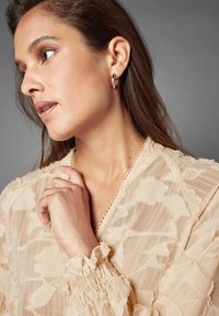 Gennemsigtig beige bluse med blomstret teksturmønster, smocked ærmer og fint blondedekoration ved halsudskæringen. Guld hoops øreringe synlige.