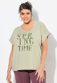 Mujer con pelo rizado, vestida con una camisa verde claro con texto de lentejuelas "SPRING TIME" y pantalones con un patrón rosa y negro, sonriendo frente a un fondo blanco.