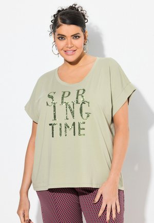 Žena s kadeřavými vlasy v světle zeleném tričku se třpytivým nápisem "SPRING TIME" a vzorovanými růžovými a černými kalhotami, usmívající se před bílým pozadím.
