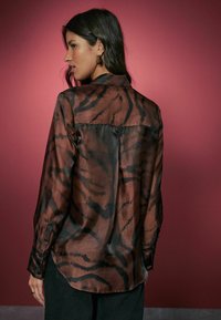 Ruda ir juoda raštuota bliuska su matinio sijono tekstūra, ilgomis rankovėmis, sagomis prie riešo ir kreivą apatinę dalį. Fonas yra vienspalvis rožinis.