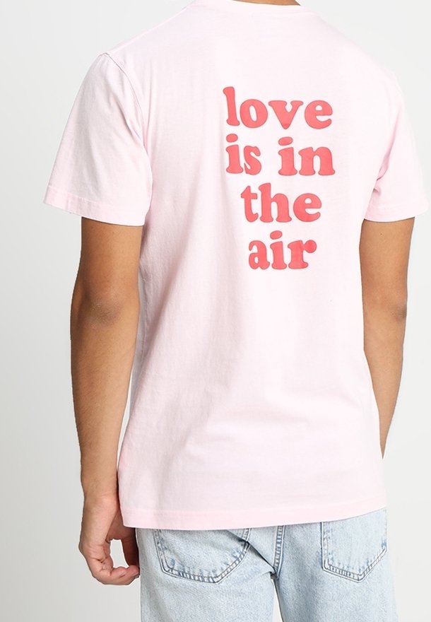Camiseta de algodón de color rosa claro con un gran texto rojo en la parte de atrás que dice "el amor está en el aire". Cuello redondo estándar y mangas cortas.