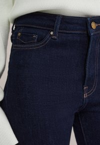 Jeans de mezclilla azul oscuro con una textura suave, que cuentan con herrajes en tono dorado, bolsillos cosidos y un diseño ajustado.