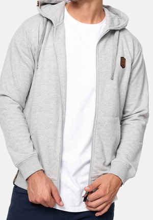 Homme portant un sweat à capuche gris clair zippé avec cordons sur un t-shirt blanc, ajustant la fermeture éclair, visible des épaules aux hanches.