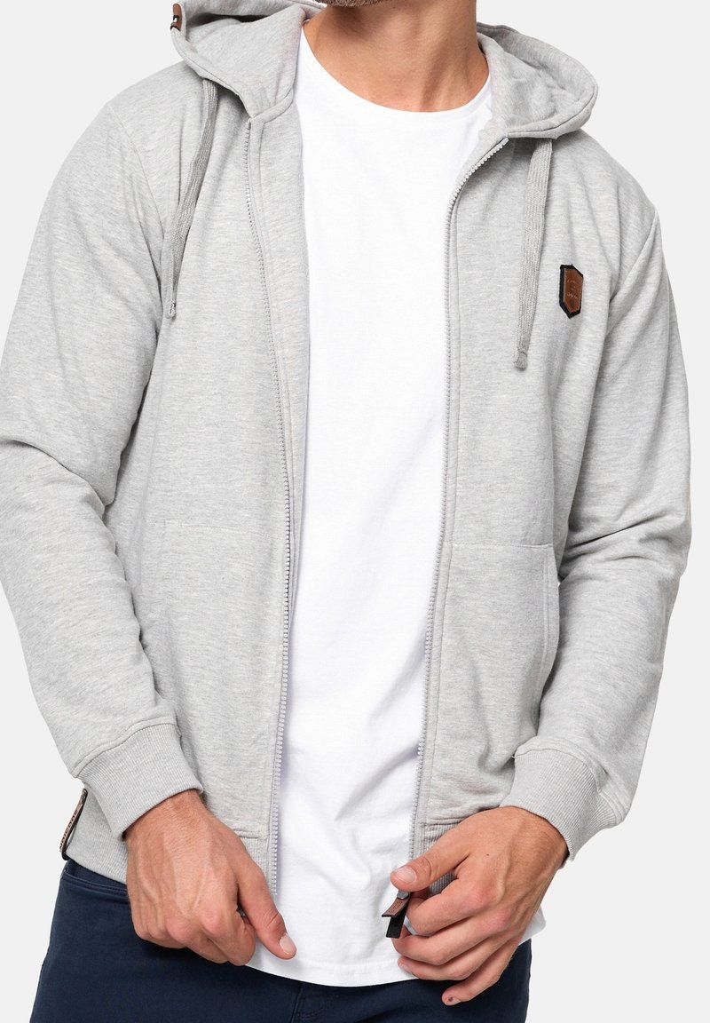 Homme portant un sweat à capuche gris clair zippé avec cordons sur un t-shirt blanc, ajustant la fermeture éclair, visible des épaules aux hanches.
