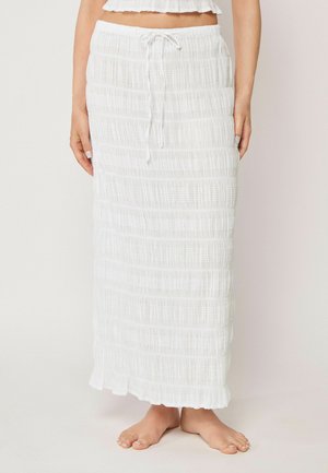 Frau trägt einen weißen, strukturierten Maxirock mit Kordelzug in der Taille und steht barfuß vor einem einfarbigen Hintergrund.