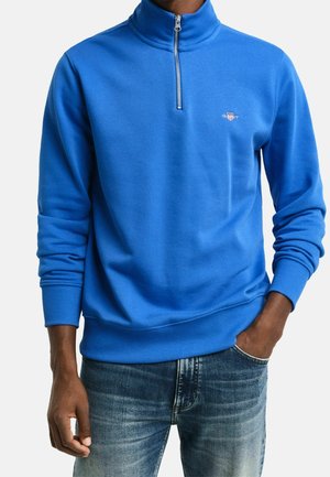 Mann trägt einen blauen Pullover mit Viertelreißverschluss, gerippten Bündchen und ausgewaschenen blauen Jeans, Hand in der Tasche.