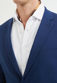 Blazer bleu marine avec revers à cran, associé à une chemise blanche à boutons ornée de fines rayures et d'un détail subtil au niveau de la poche.