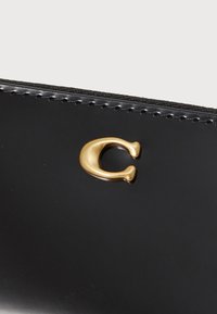 Čierna lakovaná peňaženka s výrazným zlatým logom "C". Hladký povrch s kontrastným prešívaním pozdĺž okrajov.