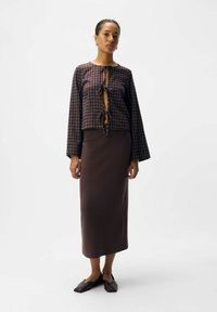Vrouw in een geruite blouse met lange mouwen en voorbindingen, bruin aansluitend midi-jurk, en platte donkerbruine schoenen, staand tegen een effen achtergrond.