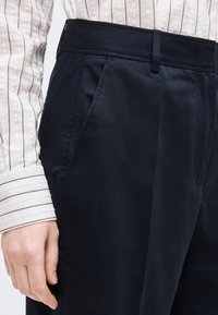 Άτομο που φοράει μαύρο ραμμένο παντελόνι και λευκό πουκάμισο με λεπτές μαύρες κάθετες ρίγες, το χέρι ακουμπάει δίπλα στην τσέπη του παντελονιού.