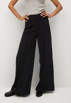 Person wearing black high-waisted wide-leg trousers, gray sleeveless top, and black shoes, standing with hands on hips against a plain background.