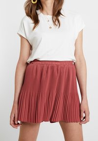 Jupe plissée de couleur rouille avec une taille élastique, associée à un t-shirt blanc à manches courtes. Design simple et texture douce.