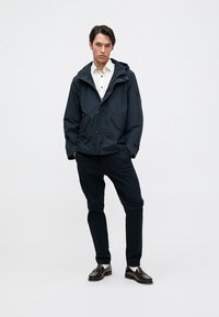 Jachetă bleumarin cu glugă, cu închidere prin fermoar și capse, având două buzunare frontale. Purtați peste o cămașă albă și asortată cu pantaloni și pantofi negri.