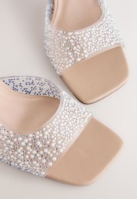 Paire de sandales plates beiges avec larges brides transparentes ornées de perles blanches et de petits clous argentés.