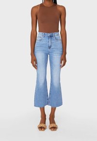 Bruin mouwloos topje, lichtblauwe high-waisted denim flare jeans met een onafgewerkte zoom, en lichtbruine gevlochten sandalen met hak, gezien van voren.