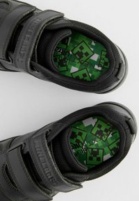 Baskets noires avec des sangles Velcro, présentant un extérieur texturé. Les semelles intérieures vertes affichent un design pixelisé avec des graphismes sur le thème de Minecraft.