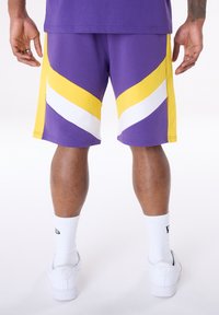 Lila sportos rövidnadrág sárga és fehér átlós cikk-cakk mintával. Az anyag könnyűnek tűnik, és fehér zoknival és cipővel van párosítva.