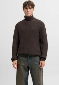 Jeune homme portant un pull à col roulé en tricot épais marron foncé et un jean noir délavé ample, se tenant devant un fond uni.