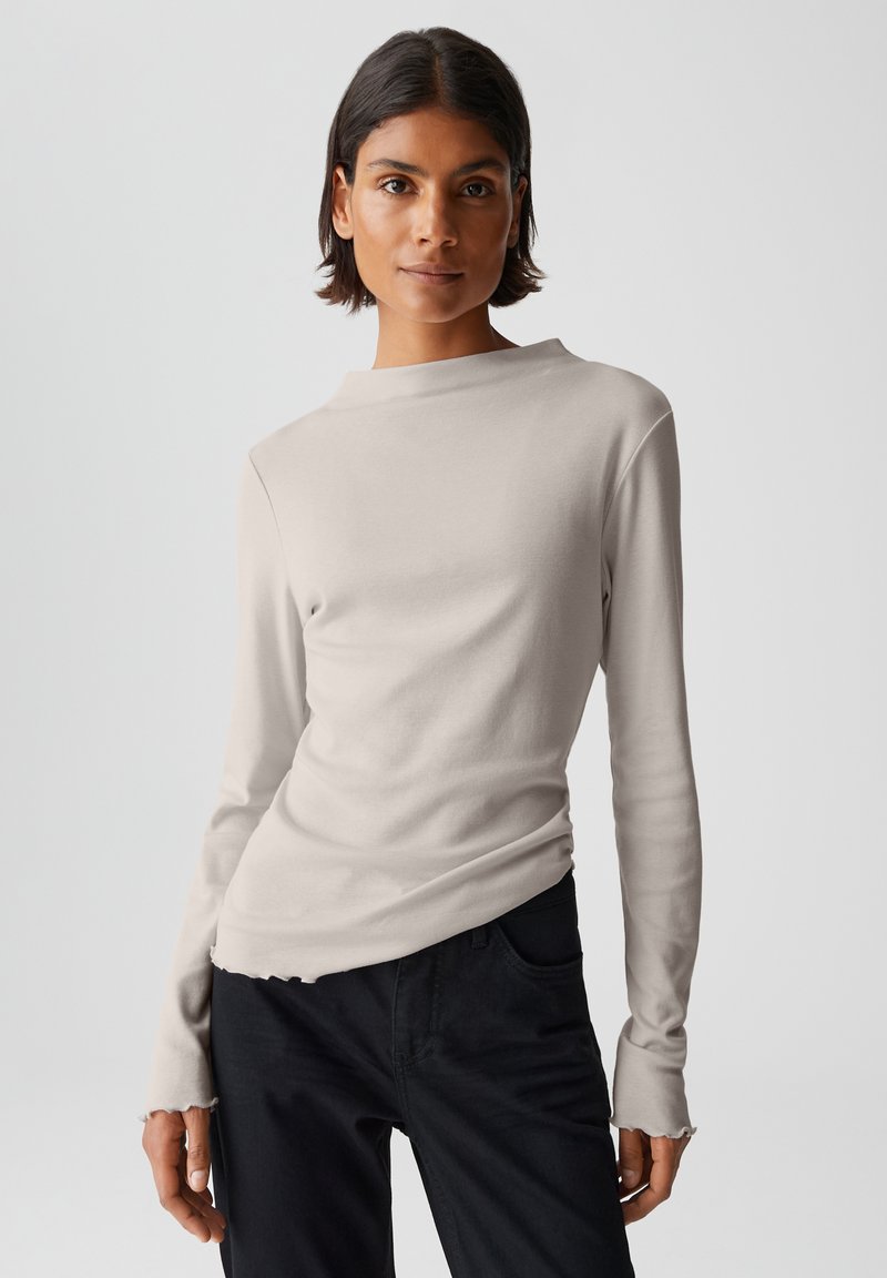 Helle beigefarbene Langarm-Bluse mit einem Boot-Ausschnitt, die eine figurbetonte Silhouette und einen leicht ausgefransten Saum aufweist. Kombiniert mit schwarzen Hosen.