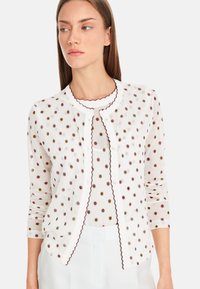 Weißer Cardigan mit runden Pünktchen in Schwarz, Maroon und Gold. Mit gewelltem Saum und vorderen Knöpfen. Lange Ärmel und leichtes Material.