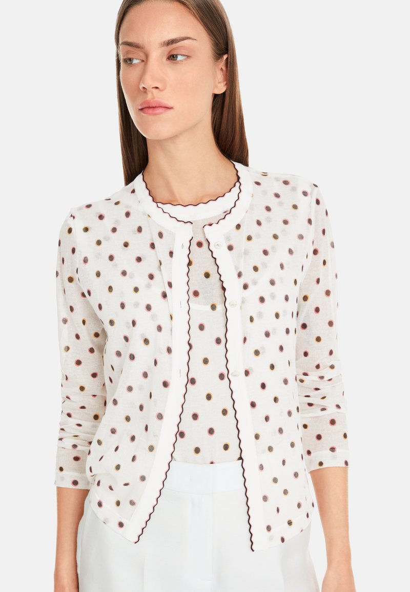 Weißer Cardigan mit runden Pünktchen in Schwarz, Maroon und Gold. Mit gewelltem Saum und vorderen Knöpfen. Lange Ärmel und leichtes Material.