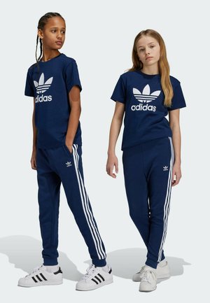 Deux enfants portant des t-shirts Adidas bleu marine et des pantalons de survêtement assortis avec des bandes blanches, debout devant un fond uni.