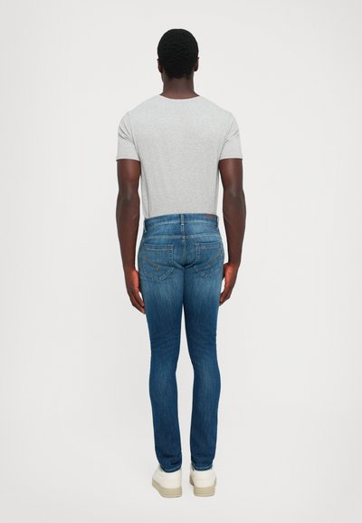 Homme debout de dos, portant un t-shirt gris clair, un jean slim bleu et des baskets blanches sur un fond uni.