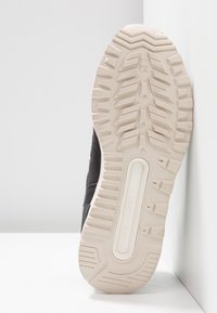 Semelle d'une chaussure New Balance noire avec un motif de bande de roulement beige reposant verticalement contre un mur d'angle blanc et le sol.