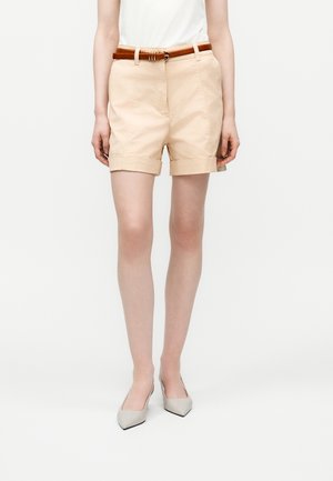 Kobieta w beżowych szortach z podwiniętymi brzegami i brązowym paskiem, białym topie bez rękawów oraz beżowych szpilkach stoi na jednolitym tle.