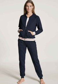 Marine blauwe zip-up jas en bijpassende joggers met geribbelde roze en witte manchetten. Glad, rekbaar materiaal; relaxte pasvorm en zijzakken.