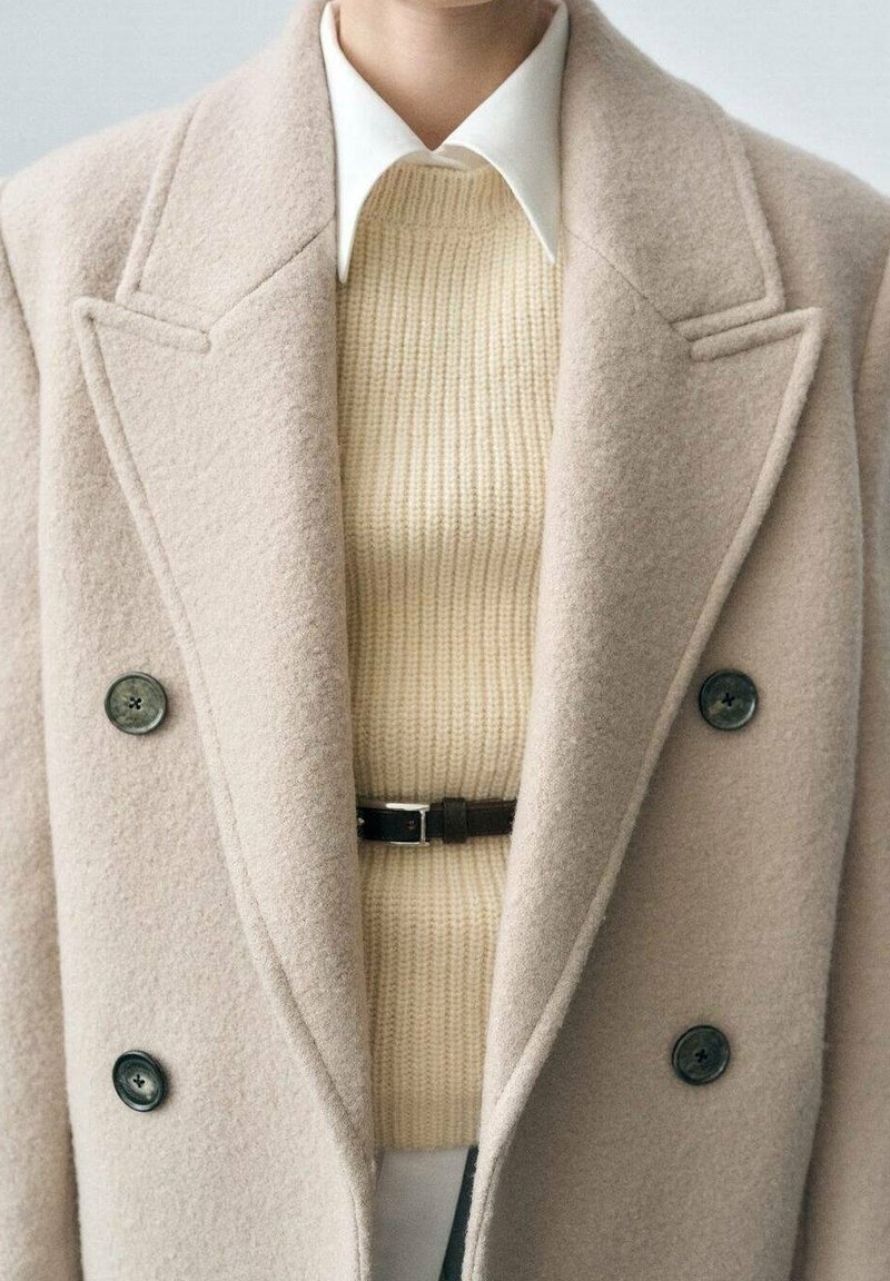 Personne portant un manteau croisé en laine beige, un pull côtelé crème, une chemise blanche à col et une fine ceinture noire à la taille.