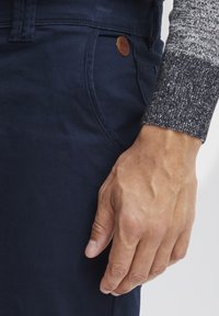 Pantalones navy con una textura suave, que cuentan con un bolsillo lateral y un acento de logo en cuero marrón. Se puede ver una manga gris texturizada.