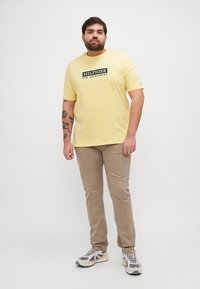 Gul bomulls t-skjorte med et sentralt svart logo og hvit tekst, kombinert med beige bukser og grå sneakers med svarte detaljer.