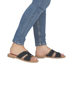 Füße in schwarzen Slip-On-Sandalen mit Nahtdetails, kombiniert mit blauen, hochgekrempelten Skinny-Jeans.