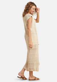 Beige gebreide maxi-jurk met een textuurpatroon en korte mouwen, met een openwerk ontwerp en zijsplitten. Draagt met bruine sandalen.