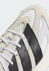 Sneakers z beżowego zamszu i siateczki, z czarnym syntetycznym wzorem trzech pasków, białymi sznurówkami i teksturowanymi akcentami. Gumowa podeszwa w zielonym odcieniu.