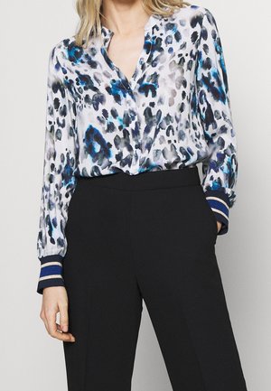 Vrouw draagt een blouse met lange mouwen en knopen, met een abstracte print in blauw en zwart, gecombineerd met zwarte hoge taillebroeken.