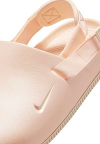 Zapato de goma rosa claro, tipo slip-on, con un upper texturizado, correa ajustable y un logo de Nike prominente en el costado.