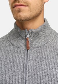 Sudadera gris con cremallera y textura acanalada, cuello alto y tirador de cremallera de cuero marrón en contraste. Vista en primer plano del escote.