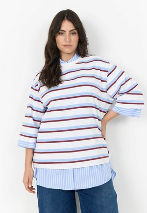 Frau mit dunklem Haar, die ein lockeres weißes Oberteil mit blauen und braunen horizontalen Streifen über einem blau-weiß gestreiften Hemd und blauen Jeans trägt.