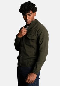 Green knitted cardigan with a small collar, featuring a chest pocket, button closure, and ribbed cuffs. Worn over a white shirt.