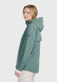Veste imperméable verte avec capuche, dotée d'une ouverture latérale, de poignets élastiques et d'un logo noir sur le bras gauche. Texture douce et lisse.