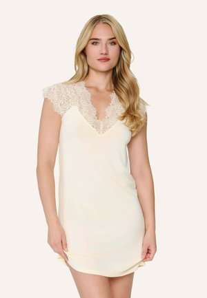 Lyshåret kvinde iført en cremefarvet, ærmeløs kjole med blonder ved skuldre og halsudskæring, stående mod en ensfarvet baggrund.