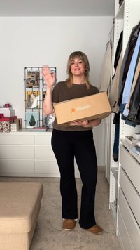 Une boîte en carton Zalando tenue par une personne portant une chemise marron et un pantalon noir, dans une pièce avec des meubles blancs et des magazines exposés.